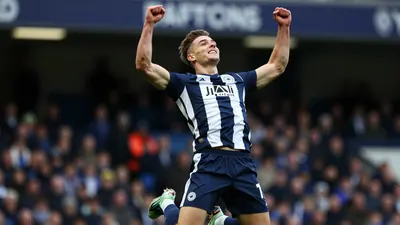 A muscular young male footballer in Derby County's navy blue and white striped kit, celebrating a goal with arms raised...