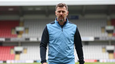 A concerned, mid-30s Derby County coach with a rugged build, wearing a plain sky blue and black training top, stands...