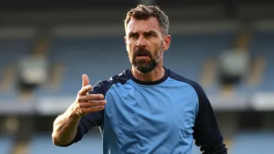 A close-up shot of a 35-year-old, ruggedly-built, bearded assistant coach, dressed in a plain, unmarked sky blue and...