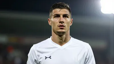 A 25-year-old Albanian footballer with a lean build and short dark hair, wearing a solid white kit made of smooth fabric...