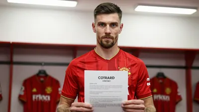 A Derby County player in a red shirt with gold trim, mid-30s, standing in the dressing room with a contract document in hand