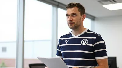 A Derby County coaching staff member in a navy blue and white striped shirt, mid-40s, standing in an office with a large...