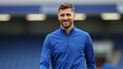 A mid-30s Derby County staff member in a solid blue training top, waist-up, smiling confidently;