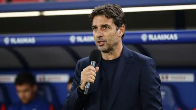 A 45-year-old male football manager with short dark hair in a navy blue suit, standing in a stadium dugout with a...